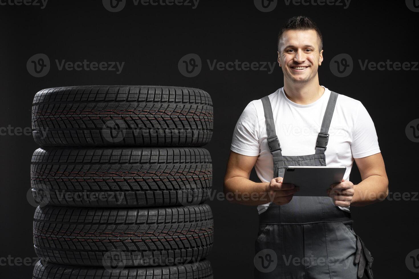 Mechanic holding tablet and smiling near stack of winter tires on black background photo