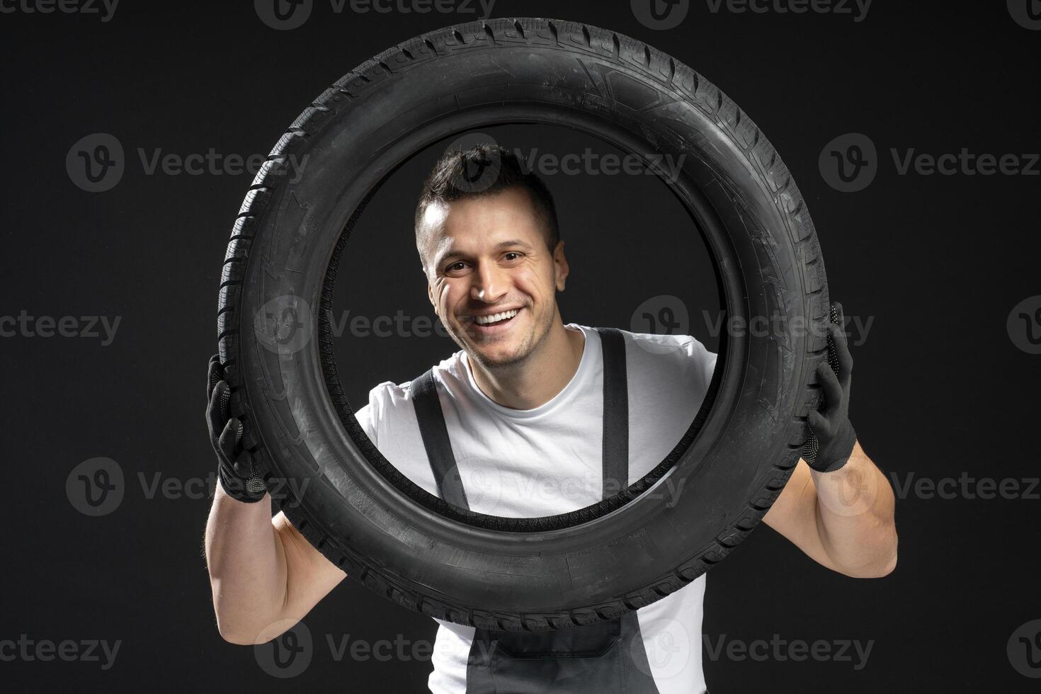 Smiling mechanic holding a tire in front of his body on black background photo