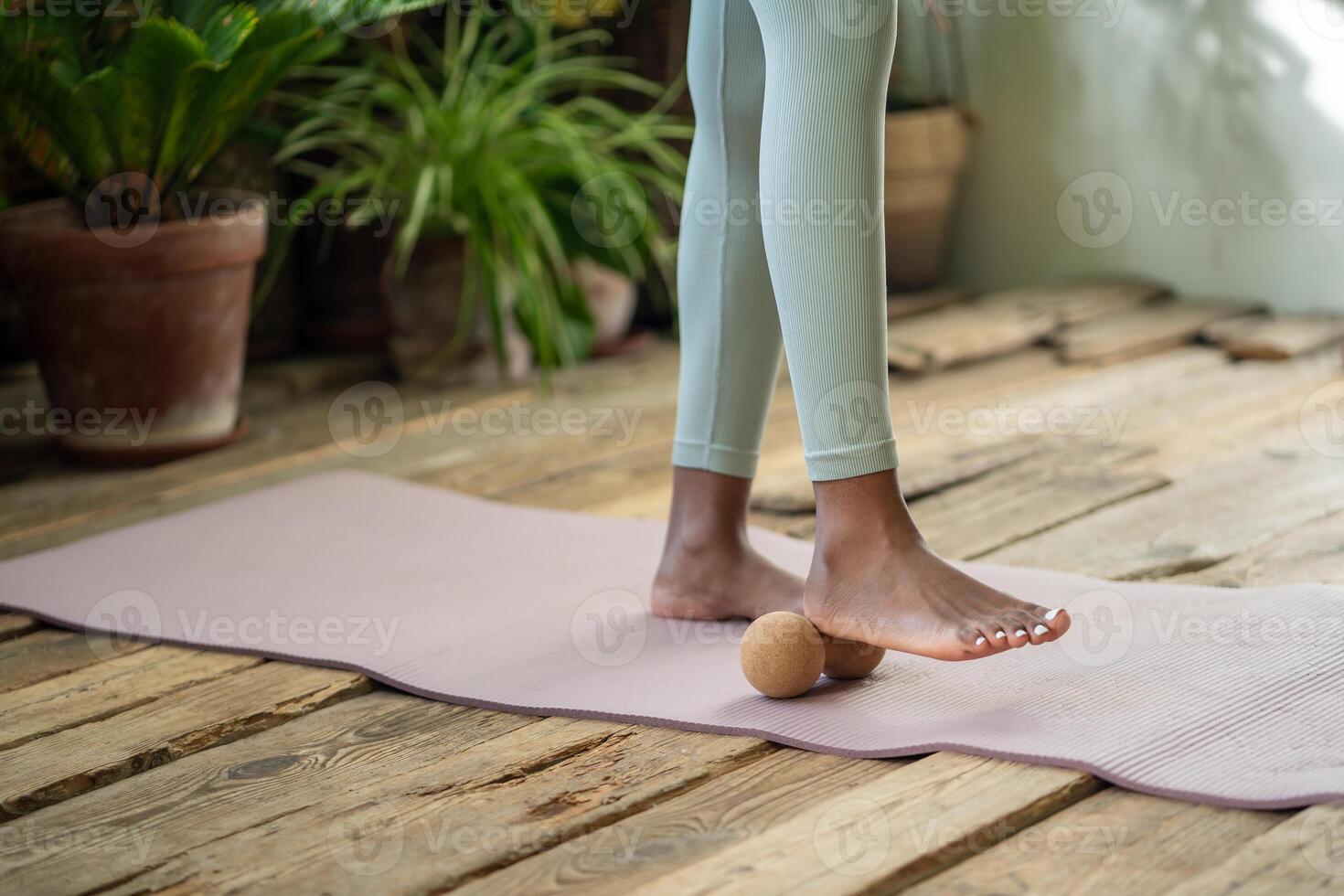 African woman stands on gym mat using massage ball to roll out tension feet spend time for self-care photo