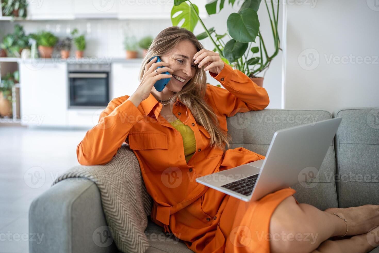 Contented woman uses laptop, talks on smartphone, smiling, laughing, making phone call sits on sofa. photo
