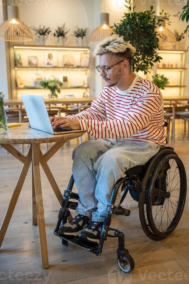 Focused disabled man in wheelchair works on laptop at cafe table. Freelancer working remotely online photo