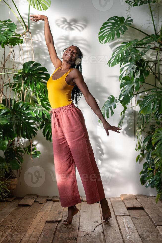 Carefree black woman dances barefoot while listening to music on headphones embraces moment indoor. photo