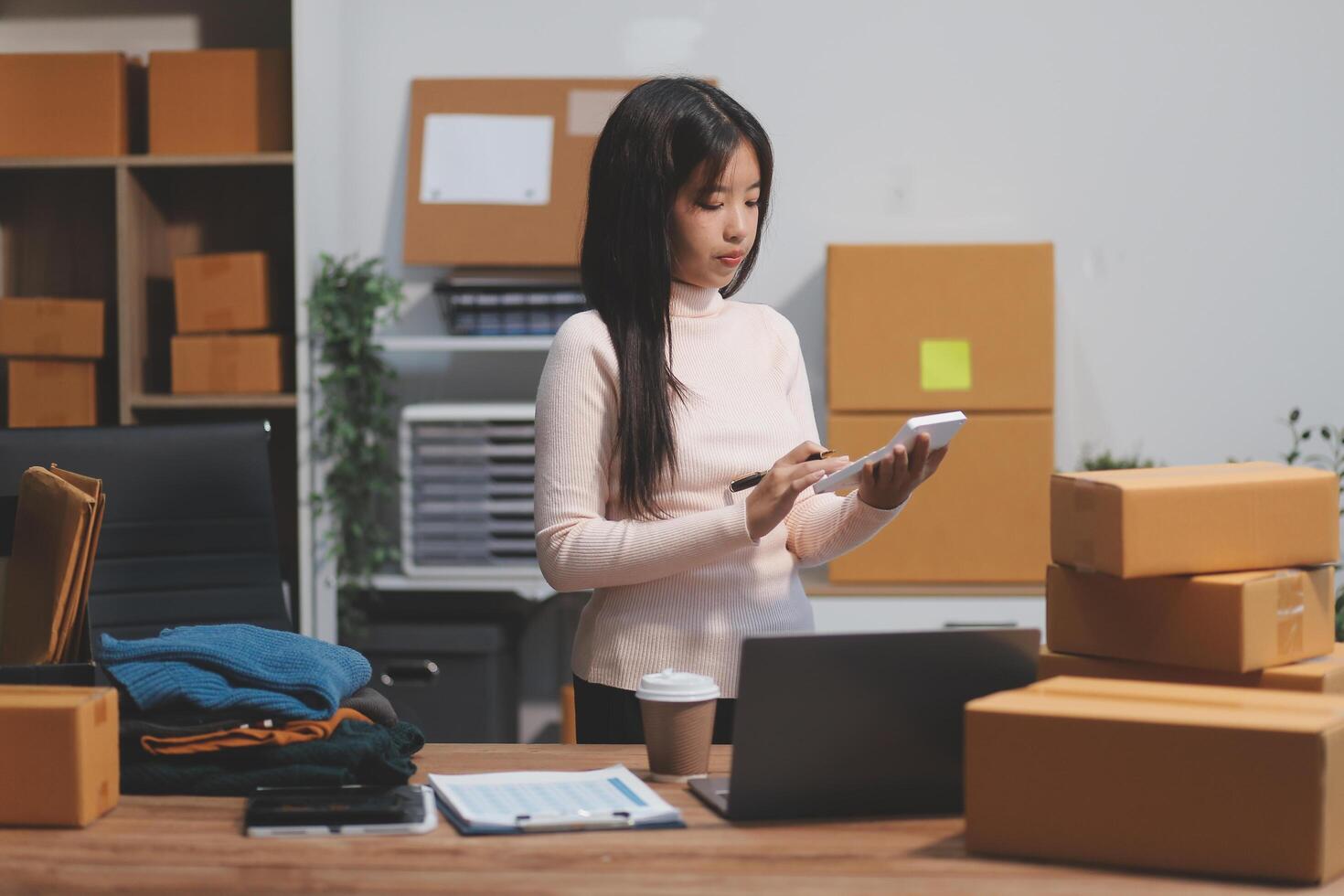 Startup SME small business entrepreneur of freelance Asian woman wearing apron using laptop and box to receive and review orders online to prepare to pack sell to customers, online sme business ideas. photo