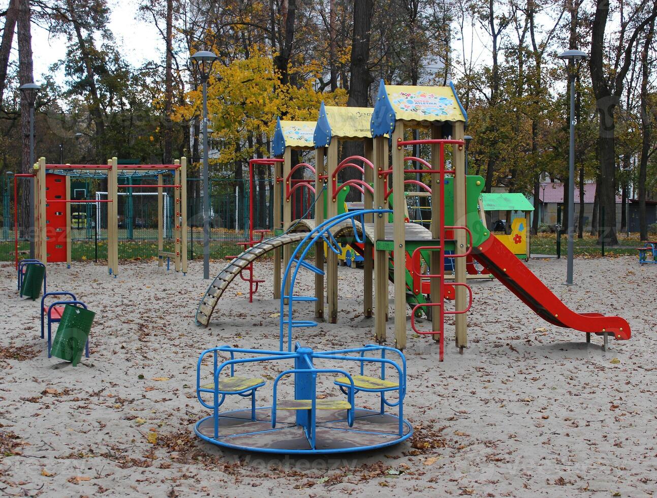 Sand covered with autumn leaves on the empty multi-colored playground with slides and small merry go round. No children at cold season at the play yard photo