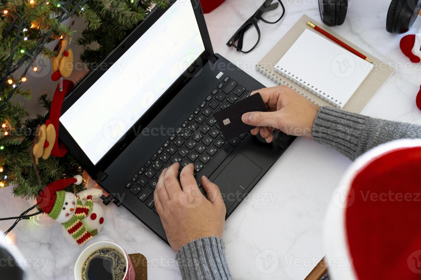Christmas online shopping. Purchase of goods and gifts for the new year via the Internet. Write a list and hold a credit card. Work desk with laptop and holiday decorations. Man's hands. Copy space. photo