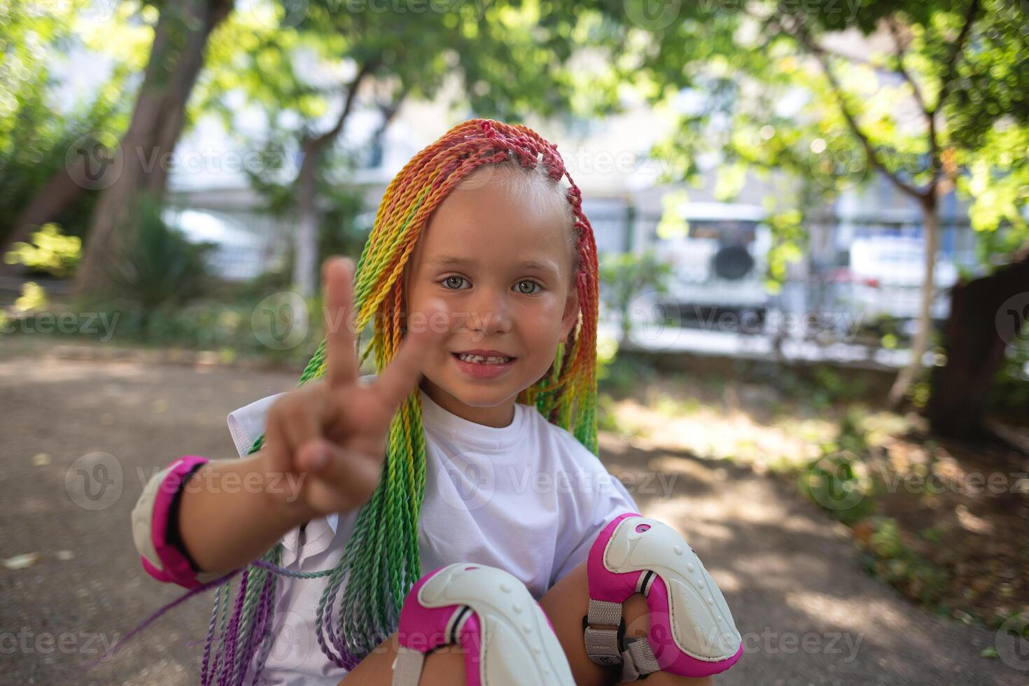 little cute girl with colorful afro pigtails sits on a skateboard and shows a peace sign photo