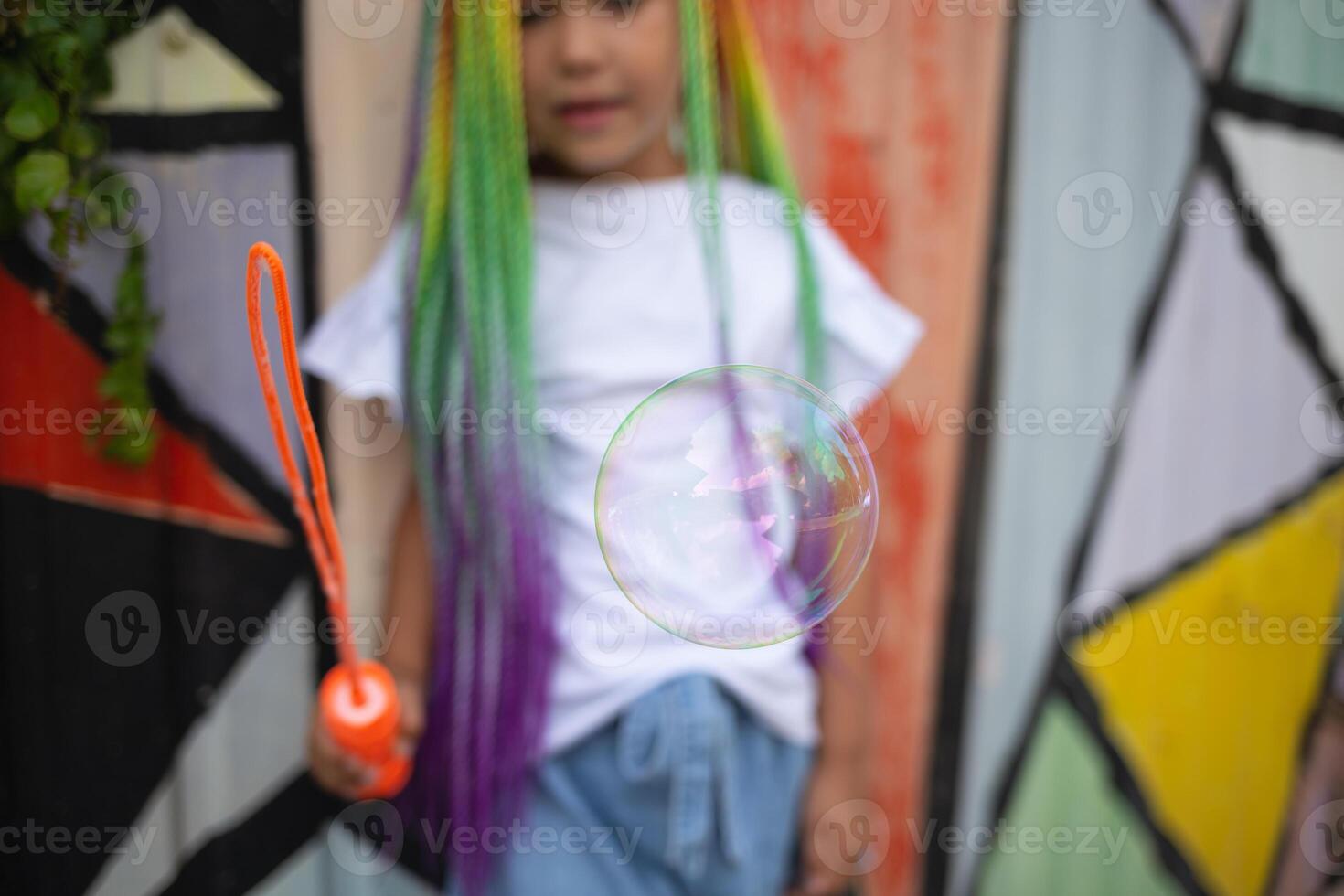 girl with multi-colored afro pigtails inflates soap bubbles against the background of a wall photo