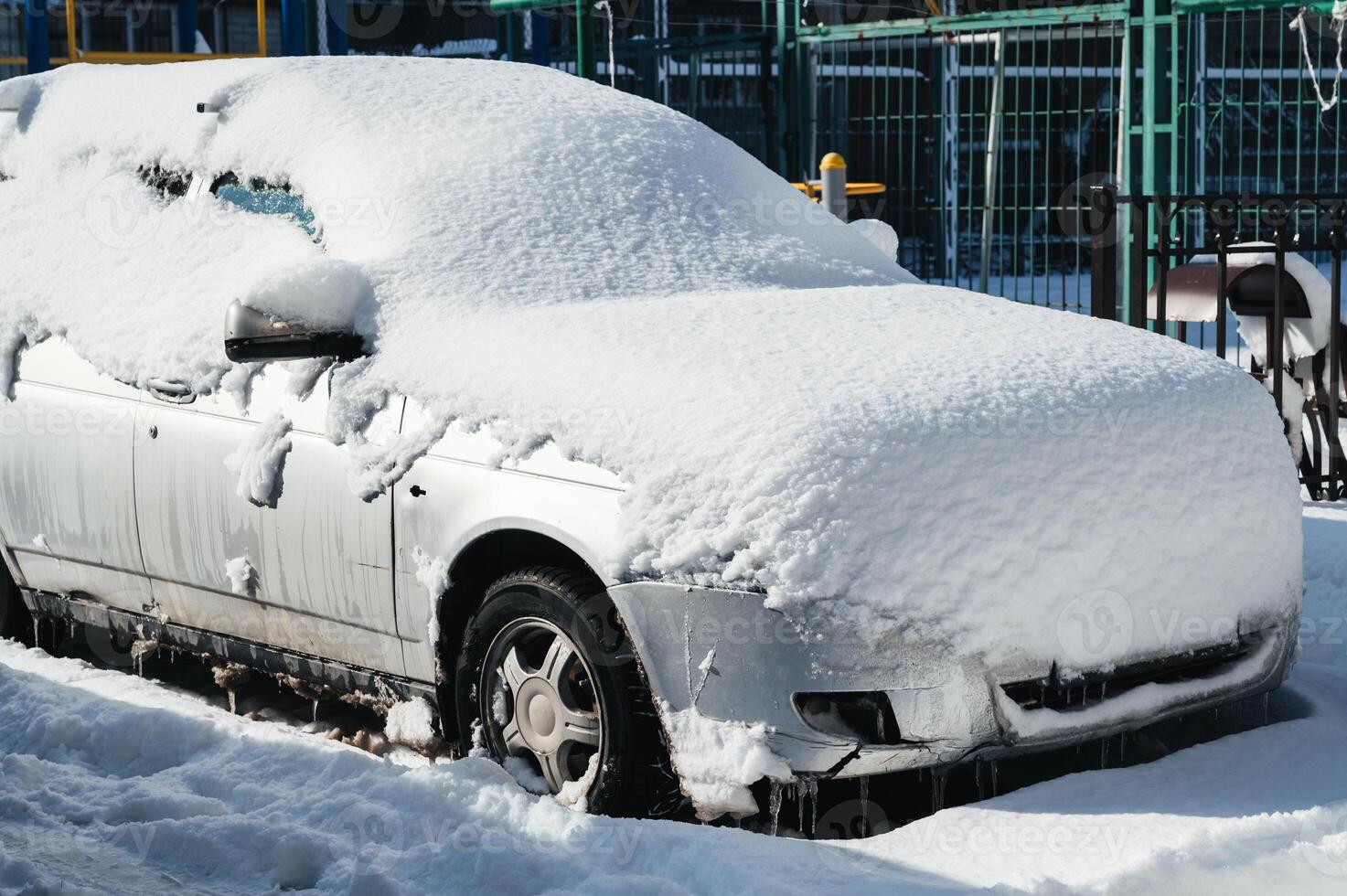 A snow-covered car is parked in the parking lot. The tires, wheels and hood of the car are covered with a white coating. photo