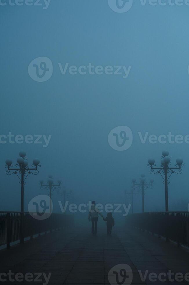 mystical silhouettes back of woman with child on a foggy bridge in the evening in autumn in fog mist photo