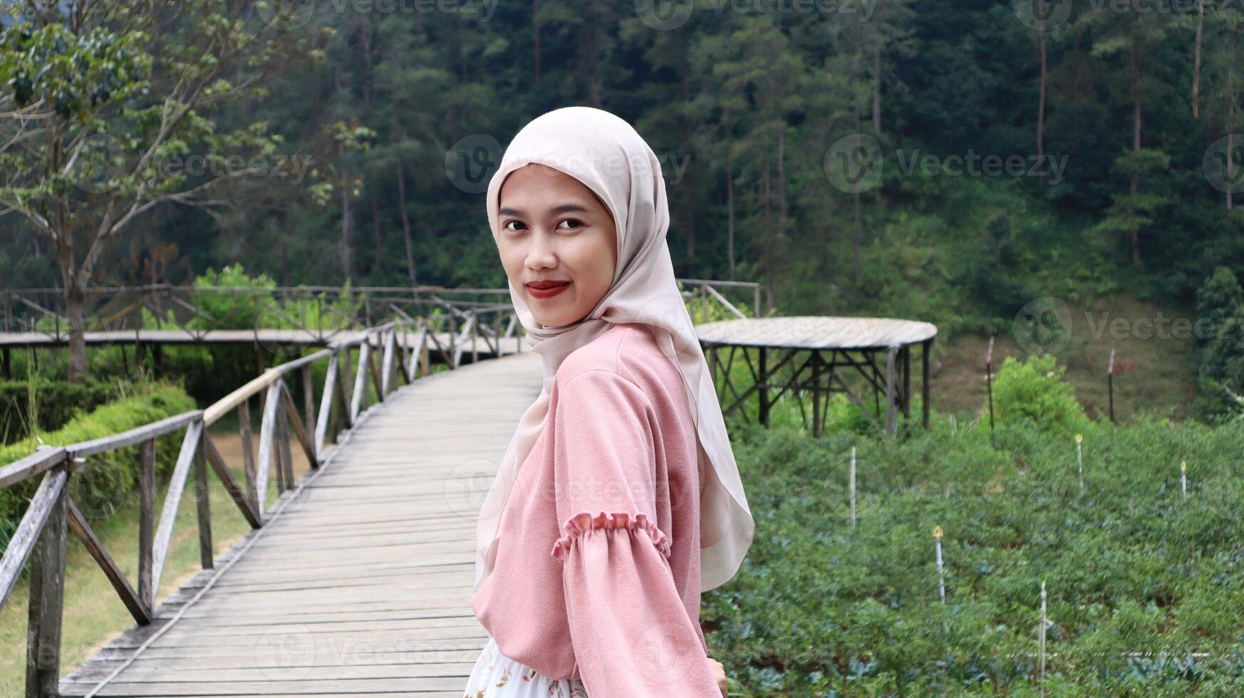 An Asian Muslim girl wearing a hijab is standing on a wooden bridge with a backdrop of natural beauty, Indonesia photo