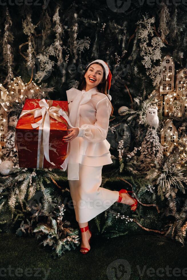 a girl in a Santa hat with a big Christmas gift in her hands on a fairy-tale island.Smiling woman in white clothes on the background of Christmas trees and small houses photo