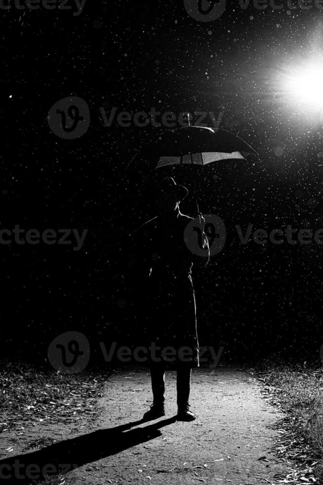 dark silhouette of a man in a raincoat and hat under an umbrella on the street in the rain photo