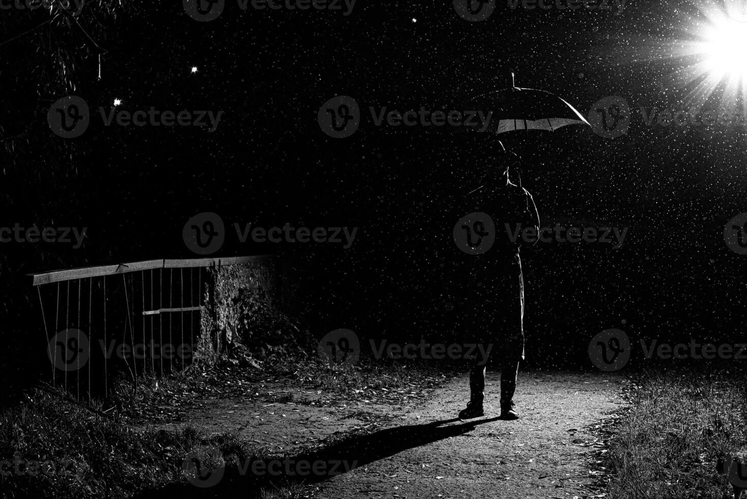 dark silhouette of a man in a raincoat and hat under an umbrella on the street in the rain photo