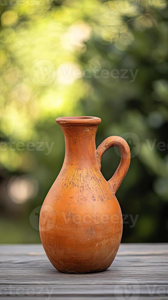 A traditional terracotta pitcher sits on a wooden table with a blurred green background. photo
