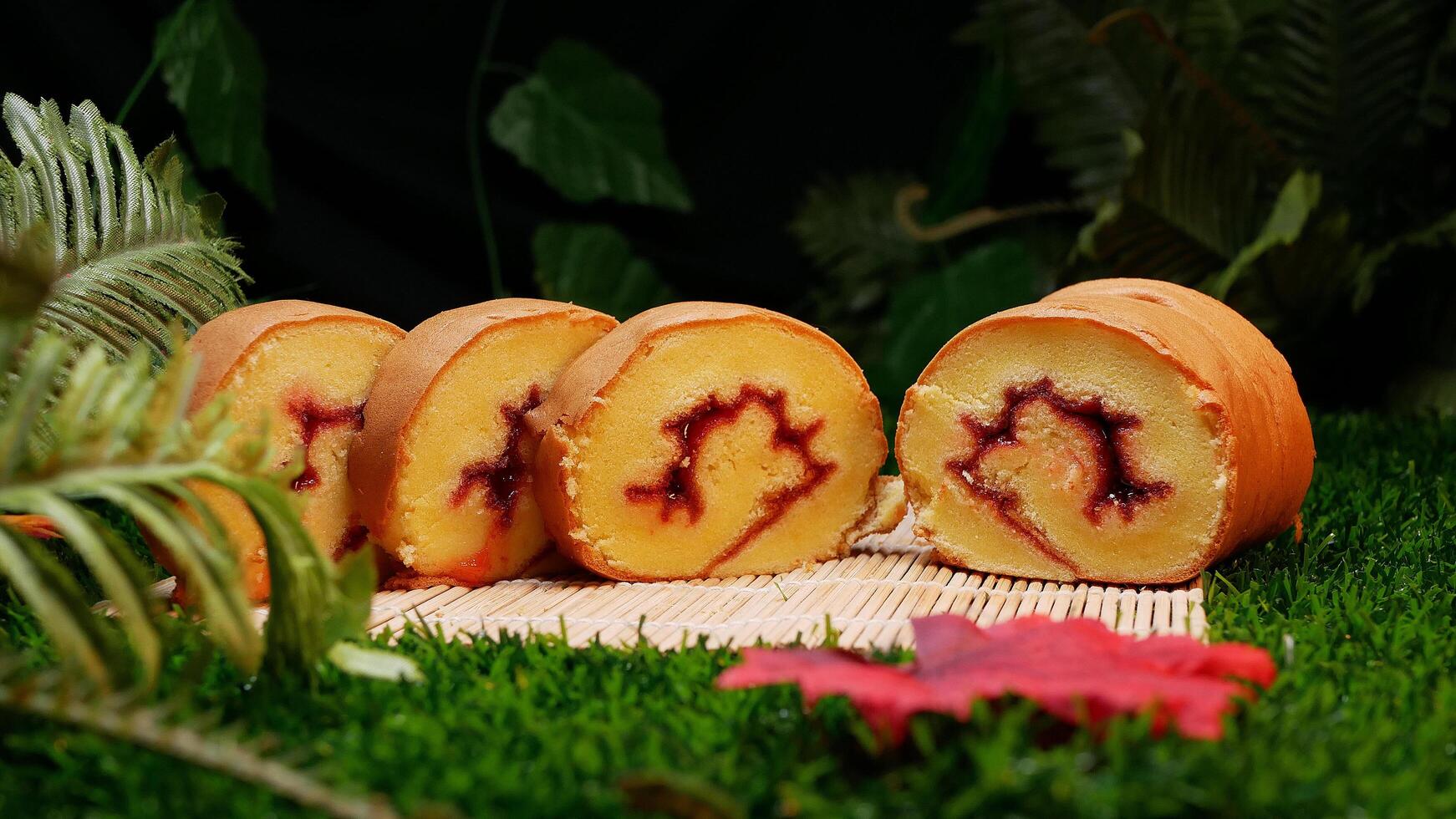 Four slices of rolled cake with red jam inside, placed on a bamboo mat and decorated with green leaves and fern plants. photo