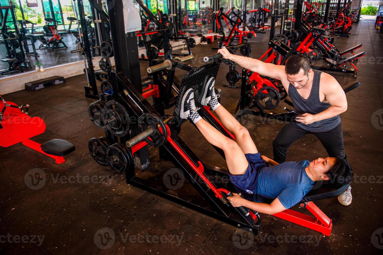 A male trainer is helping his trainee use a leg press machine by pushing the handles of the machine. photo