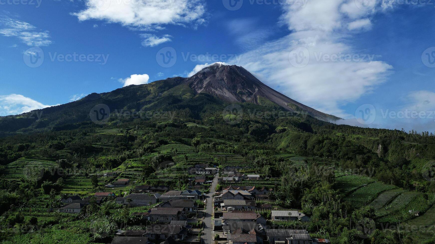 Drone view, the houses in Stabelan Village surrounded by agriculture. The majestic Mount Merapi provides a beautiful backdrop with its peak emitting wisps of thin smoke and the blue sky. photo