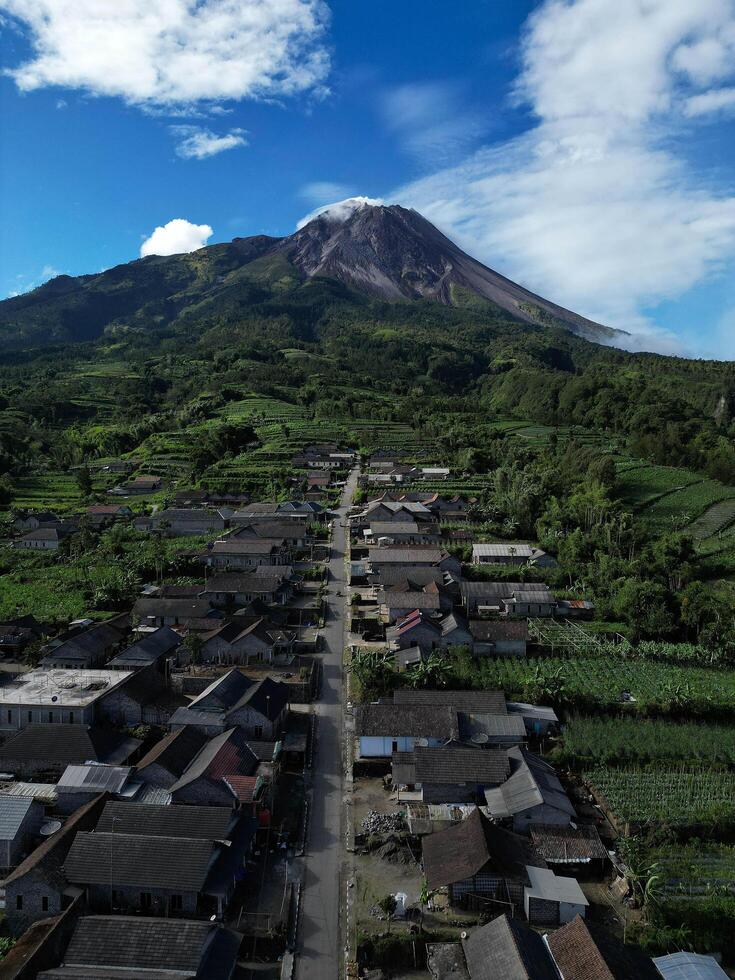 Drone view, the houses in Stabelan Village surrounded by agriculture. The majestic Mount Merapi provides a beautiful backdrop with its peak emitting wisps of thin smoke and the blue sky. photo