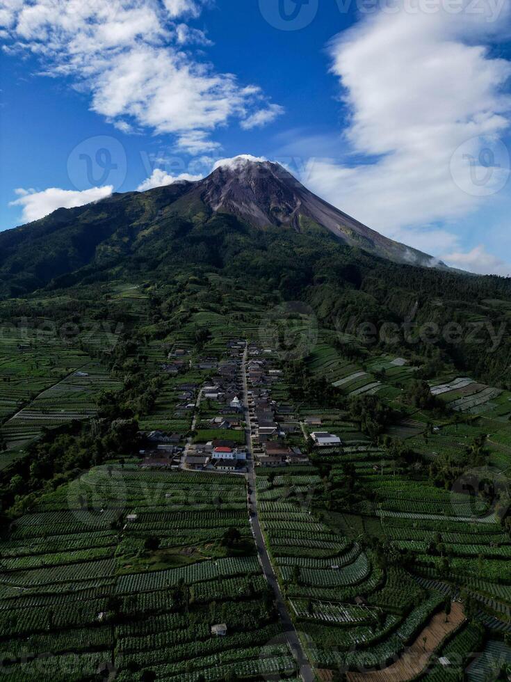 Drone view, the houses in Stabelan Village surrounded by agriculture. The majestic Mount Merapi provides a beautiful backdrop with its peak emitting wisps of thin smoke and the blue sky. photo