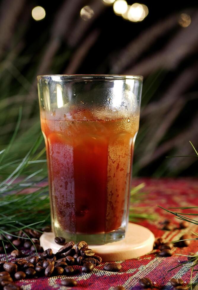 Iced Americano in a large glass with visible refreshing ice cubes, placed on a wooden coaster. Coffee beans are scattered on colorful fabric, and the background shows bokeh lights and green leaves. photo