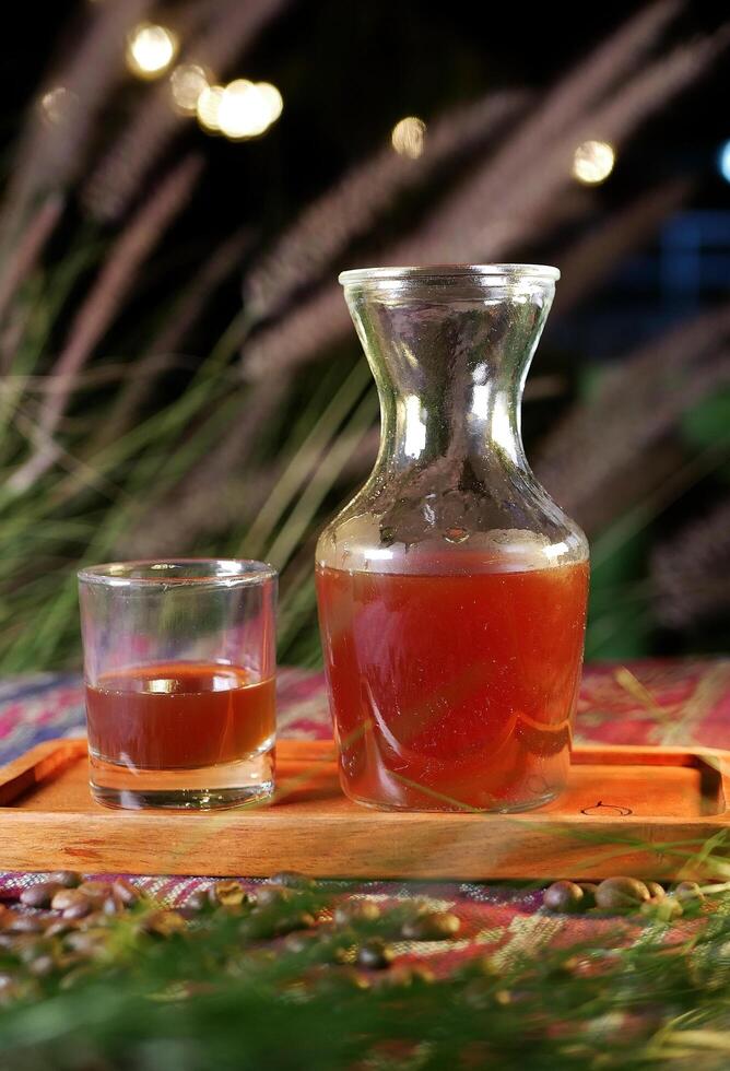 A beautifully presented Americano coffee in a clear glass and a carafe, set on a wooden tray. Scattered coffee beans, and green foliage, all illuminated by warm lights, with background bokeh lights. photo