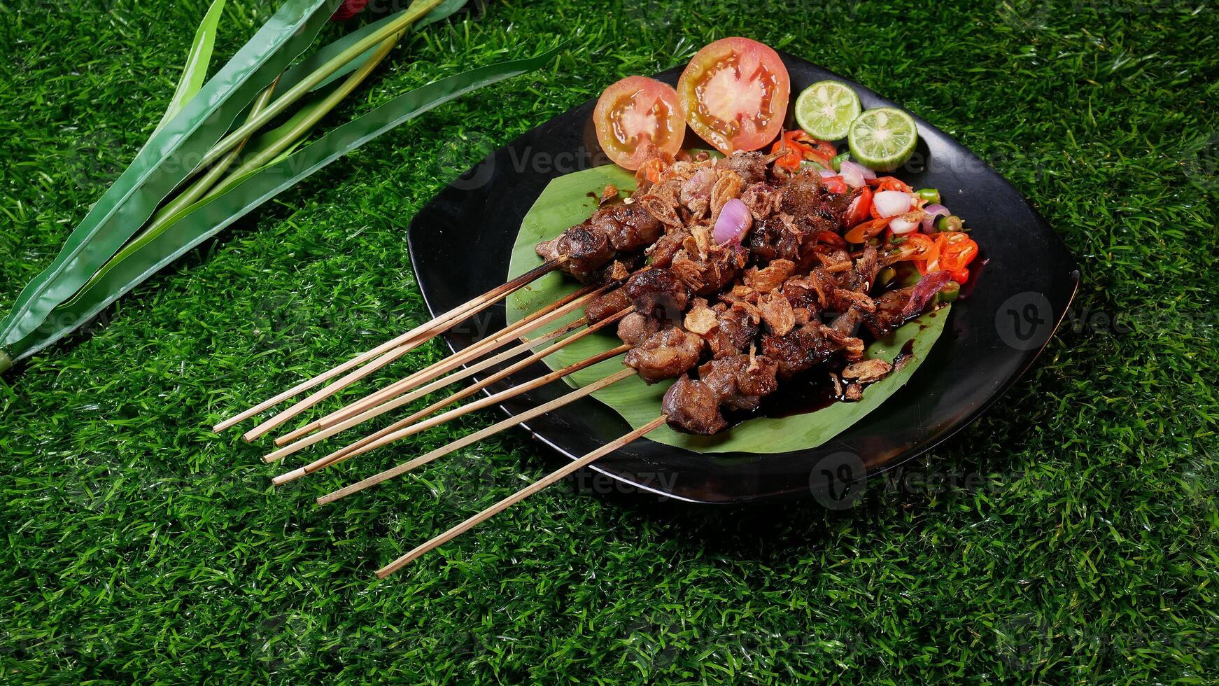 Juicy goat satay with soy sauce and fried shallots, accompanied by fresh tomato sambal, lime slices, and tomato on a banana leaf on a black plate. photo