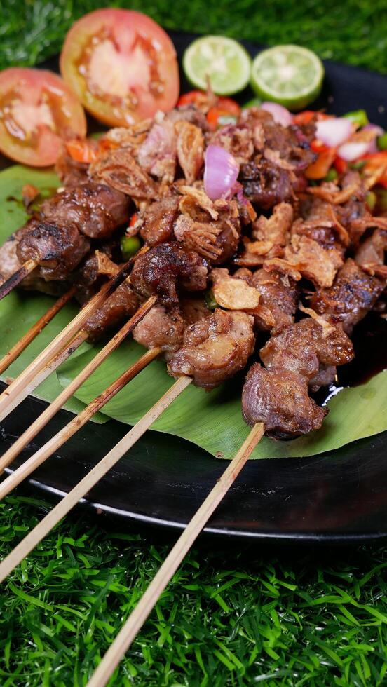 Traditional goat satay with soy sauce, sprinkled with fried shallots, served with fresh tomato sambal, lime slices, and tomato on a black plate with a banana leaf. photo