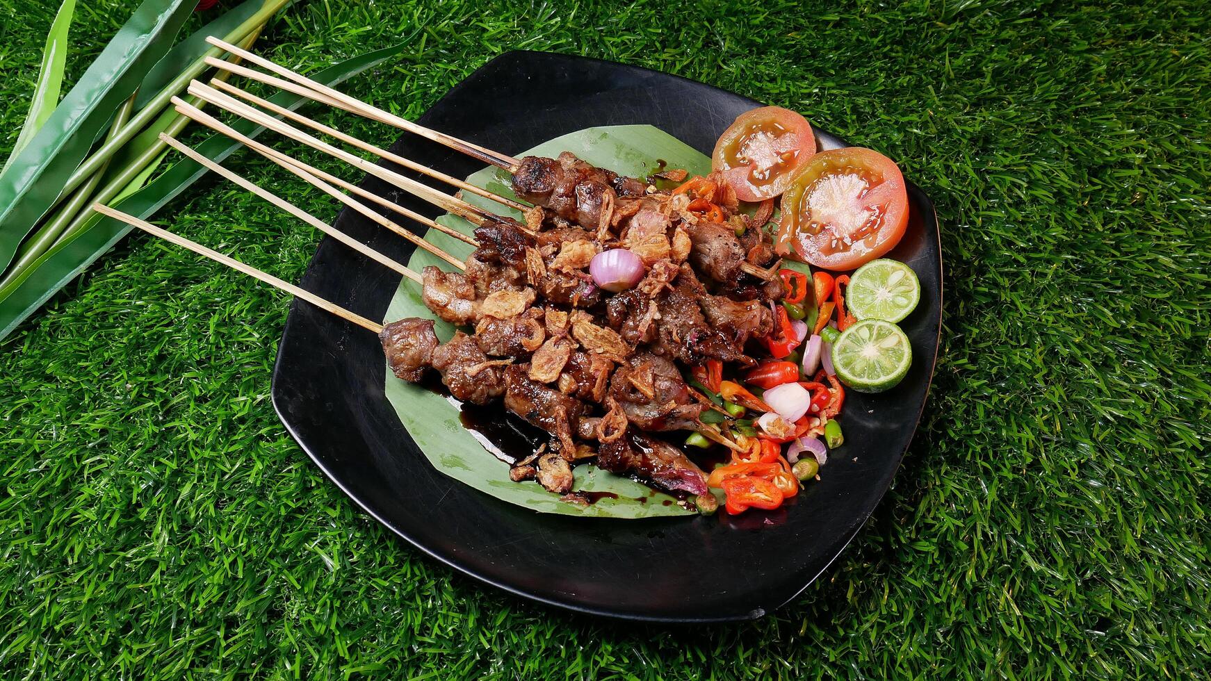 Tender goat satay with soy sauce, sprinkled with fried shallots, served with fresh tomato sambal, lime slices, and tomato on a banana leaf on a black plate. photo