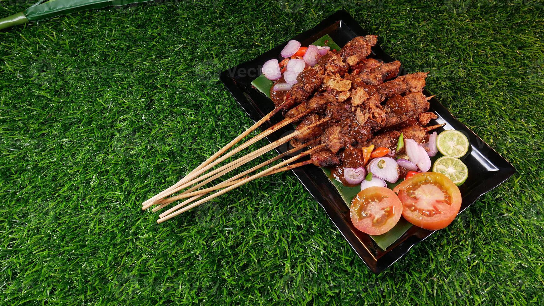 Flavorful chicken satay drenched in peanut sauce, topped with crispy fried shallots, accompanied by fresh tomatoes, shallots, chilies, and lime slices on a banana leaf over a black plate. photo