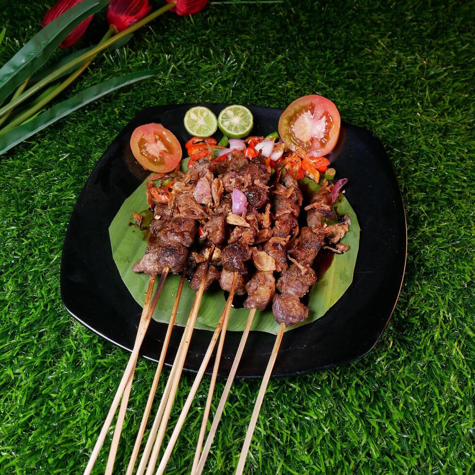 Delicious goat satay with soy sauce, garnished with fried shallots, served with fresh tomato sambal, lime slices, and tomato on a banana leaf. photo