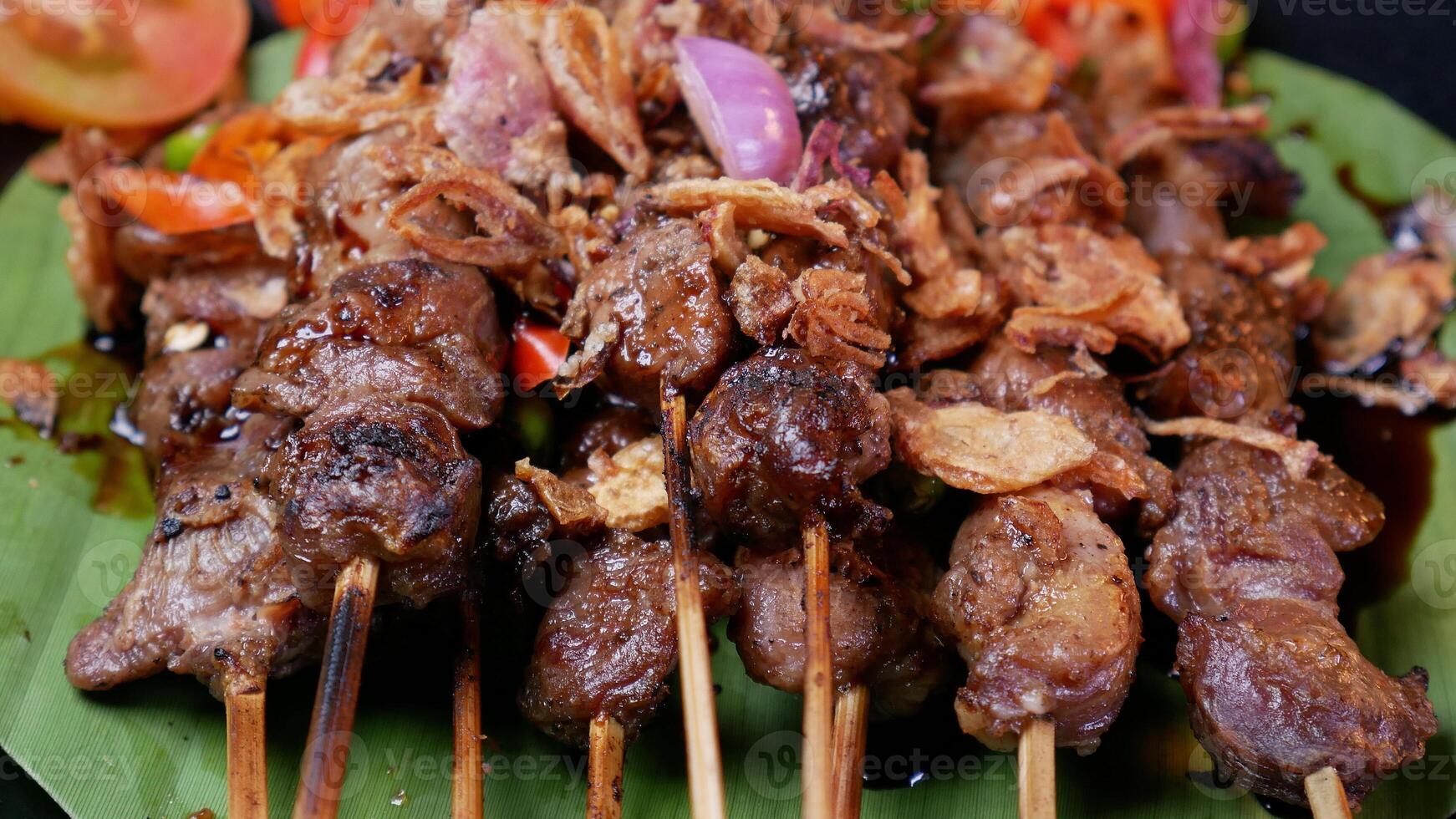 Tender goat satay with soy sauce, sprinkled with fried shallots, served with fresh tomato sambal, lime slices, and tomato on a black plate. photo