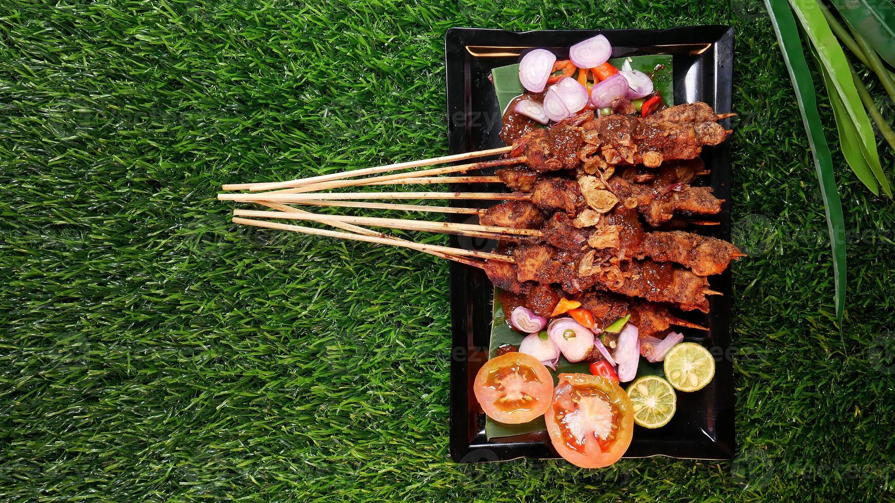 Flavorful chicken satay with peanut sauce, garnished with fried shallots, accompanied by fresh tomatoes, shallots, chilies, and lime slices on a banana leaf over a black plate. photo