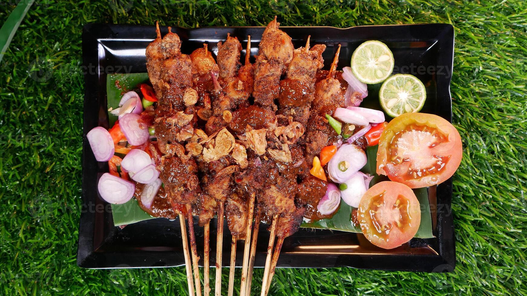 Juicy chicken satay with a generous peanut sauce, topped with fried shallots, served alongside fresh tomato slices, shallots, chilies, and lime on a black plate with a banana leaf. photo