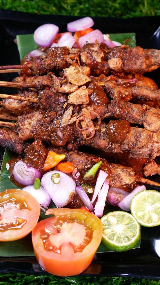 Tasty chicken satay with peanut sauce, garnished with fried shallots, served with fresh tomatoes, shallots, chilies, and lime slices on a banana leaf over a black plate. photo