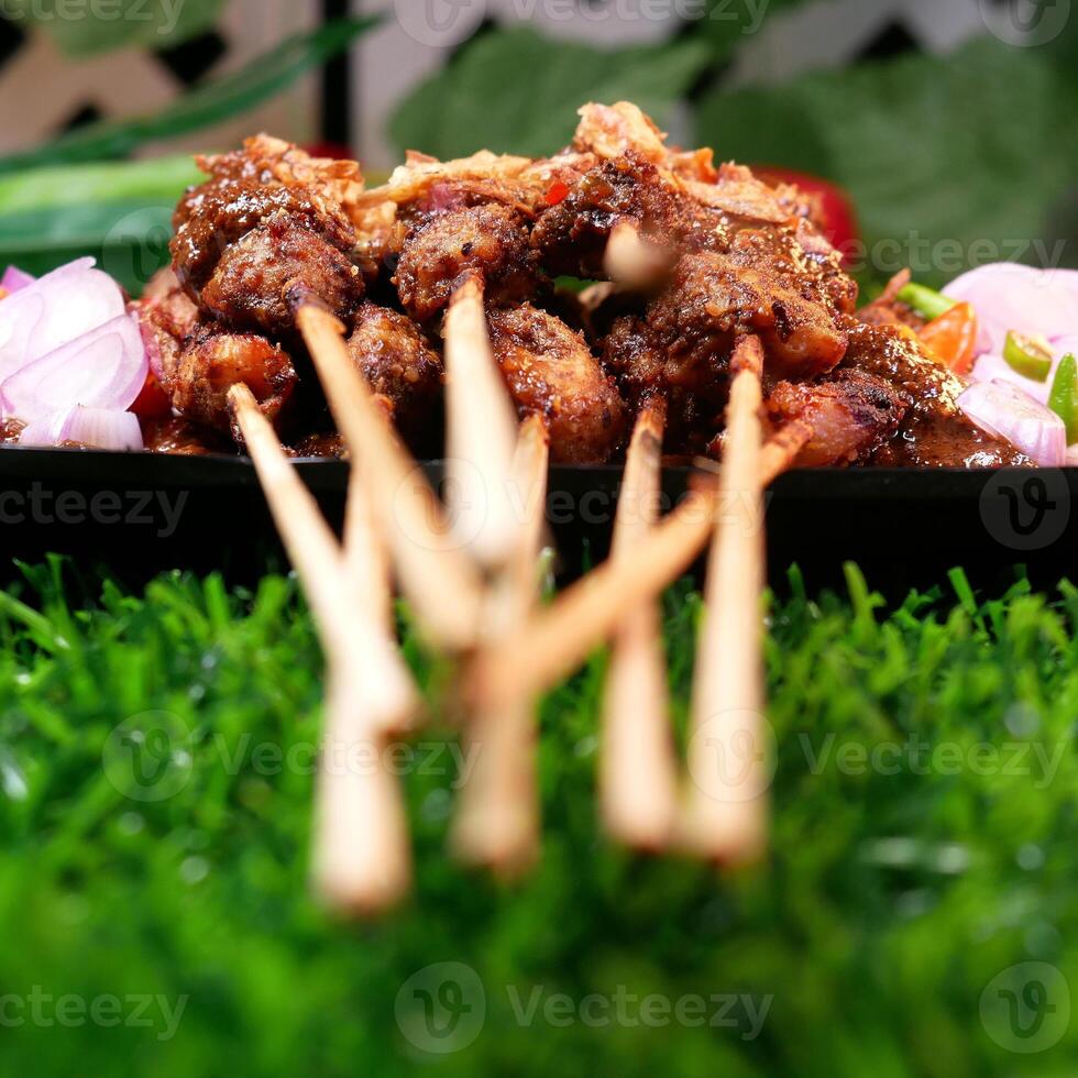 Tasty chicken satay with peanut sauce, garnished with fried shallots, served with fresh tomatoes, shallots, chilies, and lime slices on a banana leaf over a black plate. photo