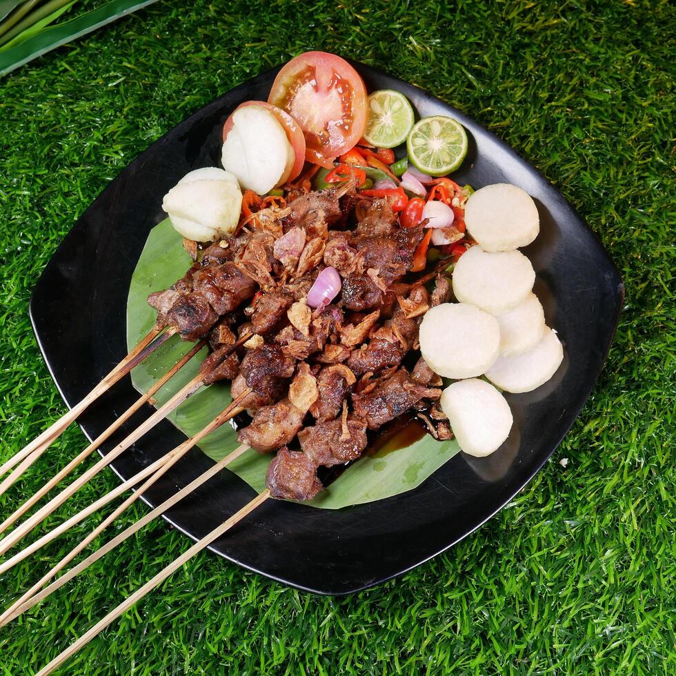 Special goat satay with peanut sauce and a sprinkle of fried shallots and red onions, served with rice cakes, tomato slices, and lime on a banana leaf. photo