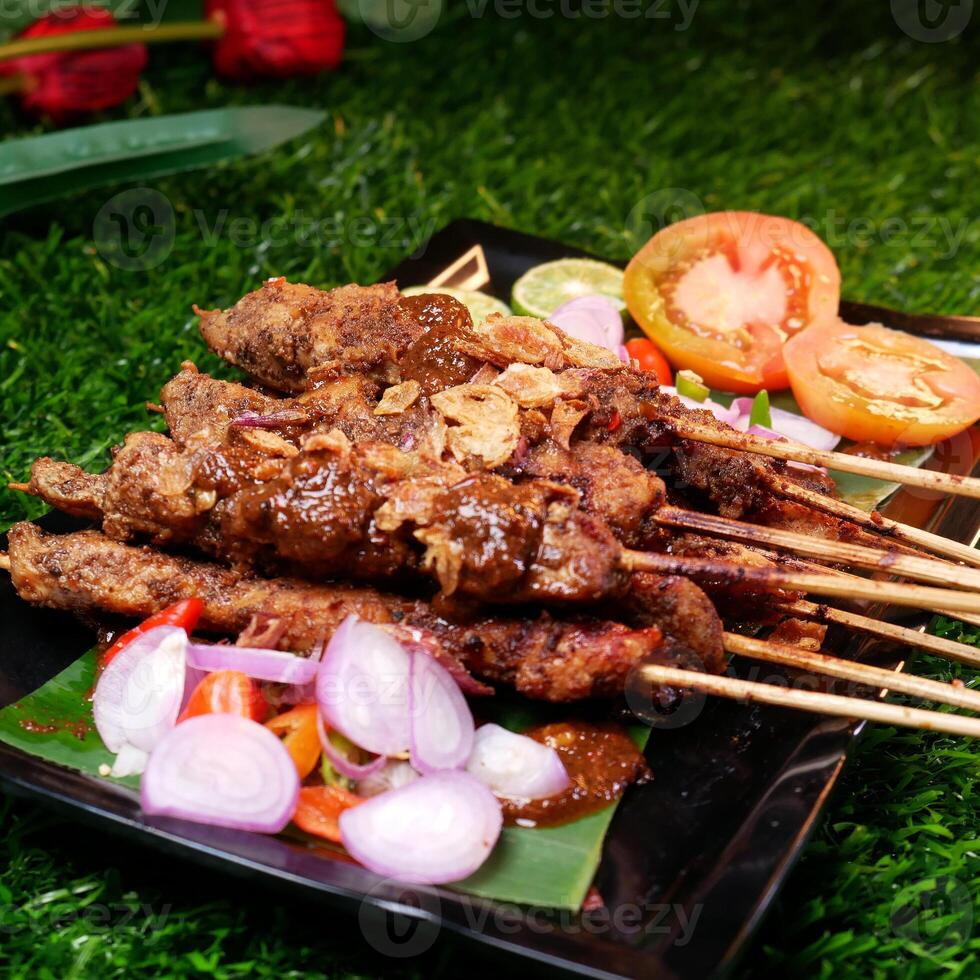 Juicy chicken satay with peanut sauce, garnished with fried shallots, and served with fresh tomato slices, shallots, chilies, and lime on a banana leaf. photo