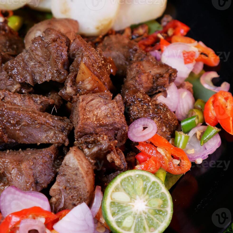 Perfectly seasoned grilled goat sprinkled with fried shallots and red onions, accompanied by rice cakes, lime slices, and fresh tomatoes on a banana leaf. photo
