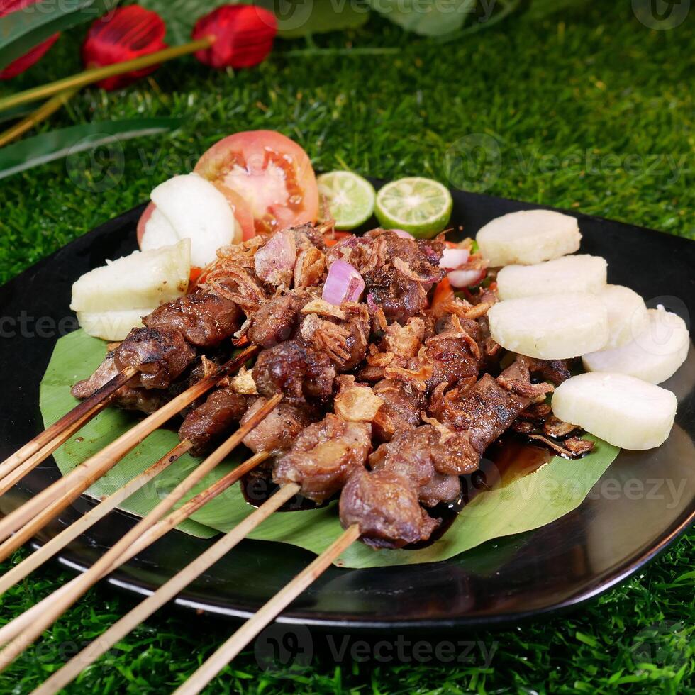 Delicious goat satay sprinkled with fried shallots and red onions, served with rice cakes, fresh tomatoes, and lime on a black plate with a banana leaf. photo