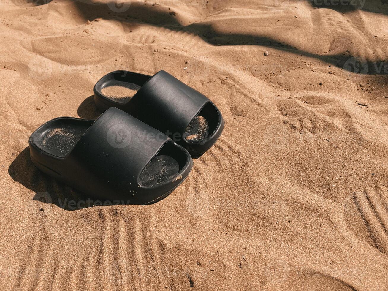 Pair of black slippers standing on soft golden sand under warm sunlight. Shadows and footprints surround scene, calm, relaxation and simplicity. Perfect for beach vacation and summer outdoor leisure photo
