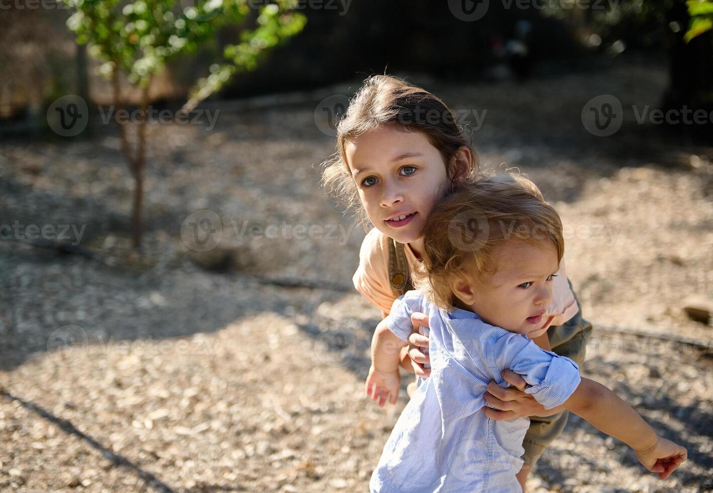 Playful siblings outdoors in a sunny park setting, an older sister lovingly embraces and supports her younger brother, capturing a moment of care, warmth, and family bonding in nature's backdrop. photo
