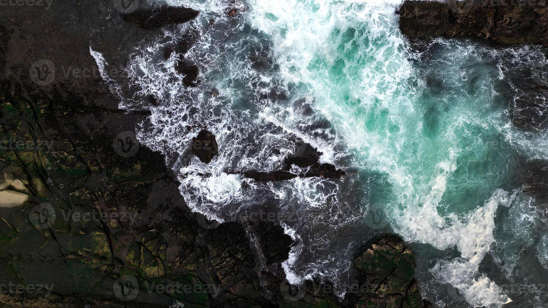 The ocean waves crash against the black rocks, creating white foam that contrasts with the dark green seawater. The water flow appears strong, breaking through the rough rock crevices. photo