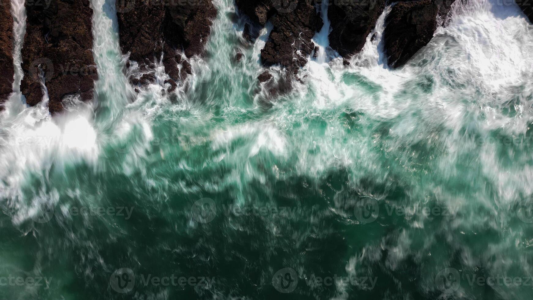 The ocean waves crash against the black rocks, creating white foam that contrasts with the dark green seawater. The water flow appears strong, breaking through the rough rock crevices. photo
