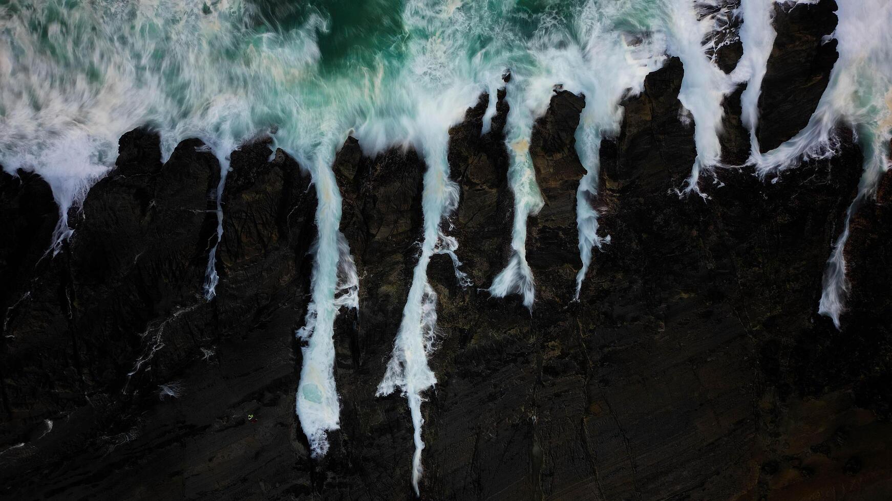 The ocean waves crash against the black rocks, creating white foam that contrasts with the dark green seawater. The water flow appears strong, breaking through the rough rock crevices. photo