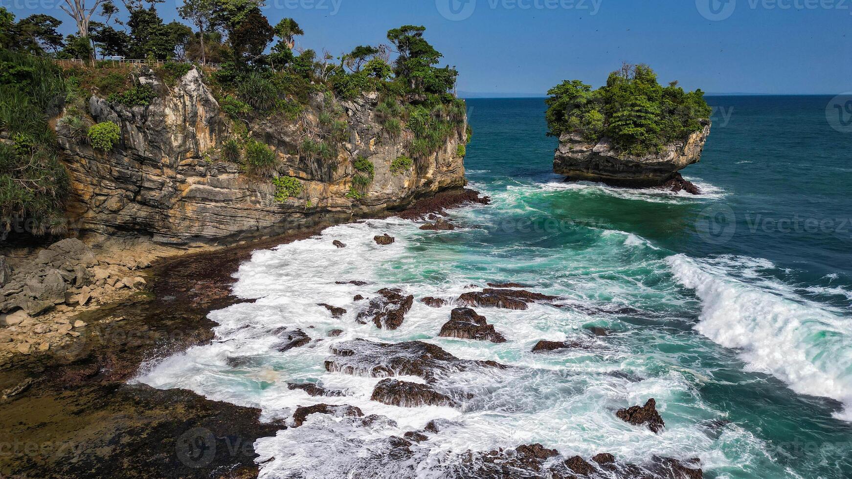 An exotic view of Karang Bokor Beach with high and green rock cliffs, surrounded by clear blue seawater. The waves crashing against the rocks create a beautiful photo