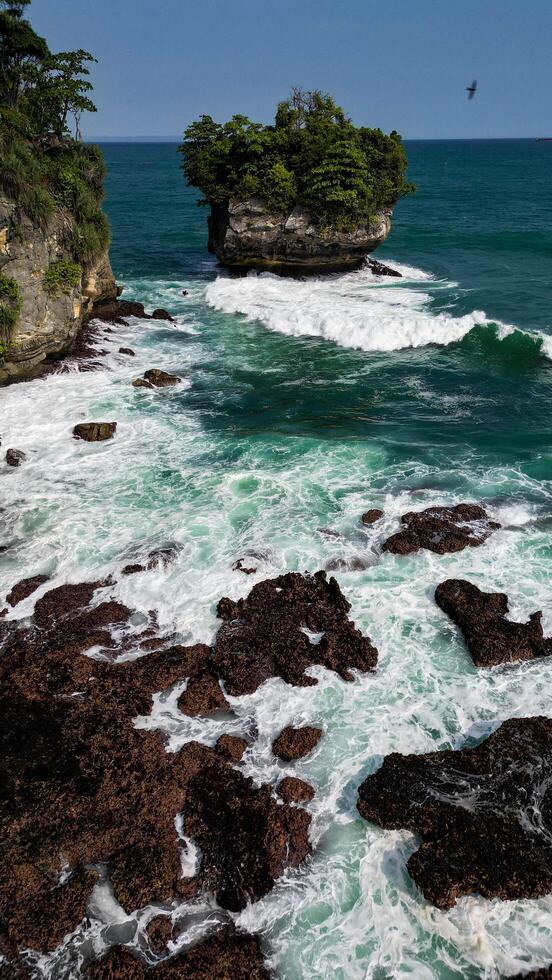 The majestic and green rock cliffs at Karang Bokor Beach stand tall amidst the vast blue sea. The white waves crashing against the rocks add a calming photo