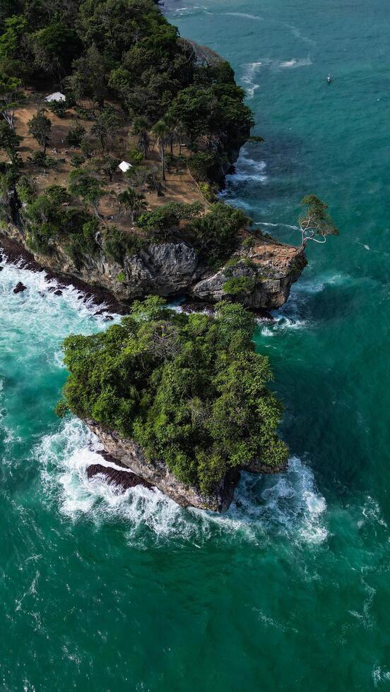 An aerial photo of Karang Bokor Beach displaying spectacular rock formations with green trees on top, surrounded by clear blue seawater and crashing waves.