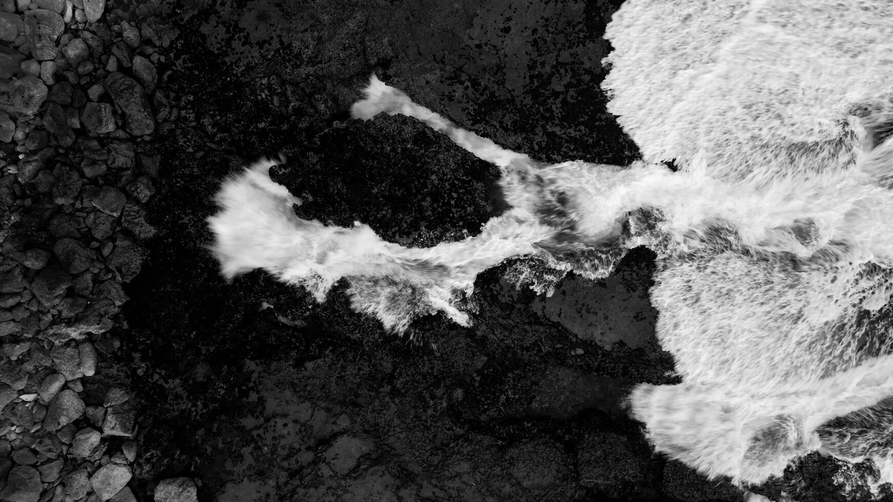 Ocean currents crash hard against the black rocks on the shore, forming white foam that contrasts with the rough, hard surface of the stones below. Sawarna Beach, Banten, Indonesia. photo