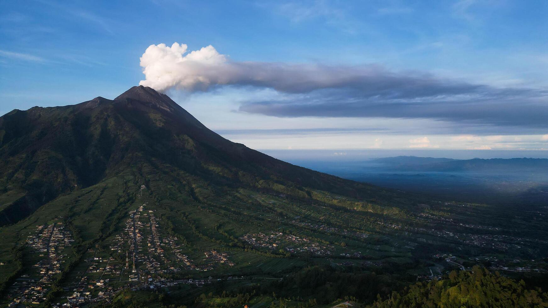 The Beauty and Majesty of the Smoking Mount Merapi. The towering mountain is surrounded by lush green valleys and scattered villages at its base. photo