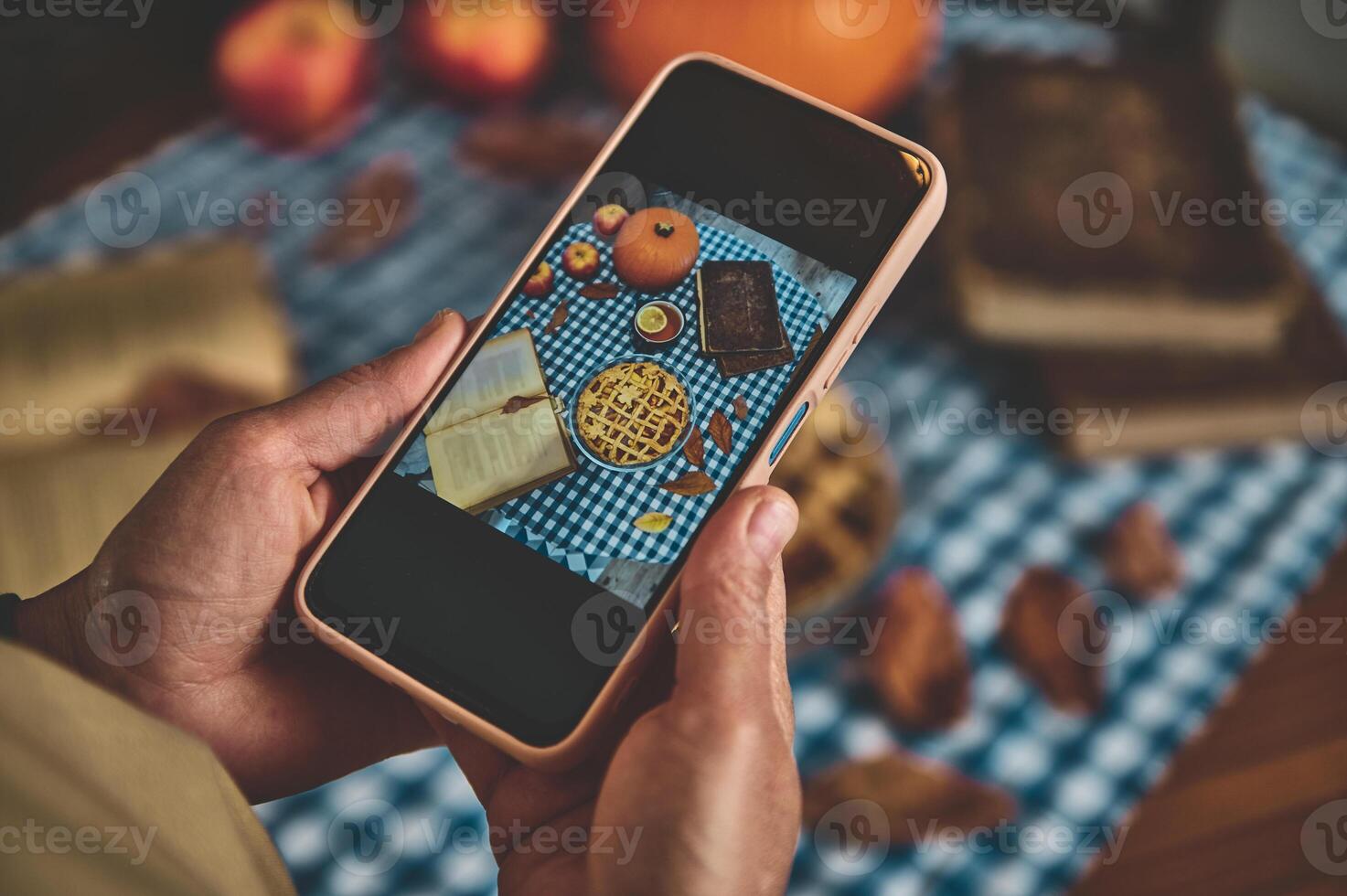 Capturing the essence of autumn with smartphone. Top-down view of a warm, cozy setup featuring an apple pie, books, and seasonal decorations on a checkered blue and white tablecloth. photo